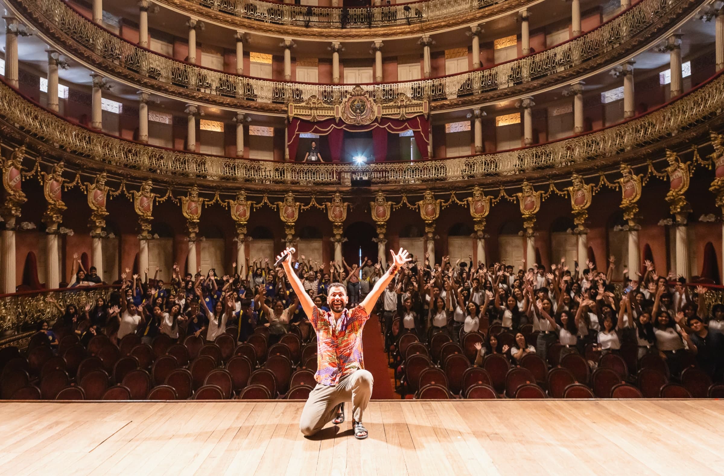 Juraci Júnior ajoelhado no palco do Teatro Amazonas, de braços erguidos, com a plateia lotada também de braços erguidos atrás dele.
